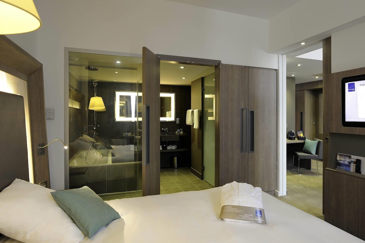 A hotel room with a bed, a bedside lamp, a mirror, a TV, and a desk with a chair.
