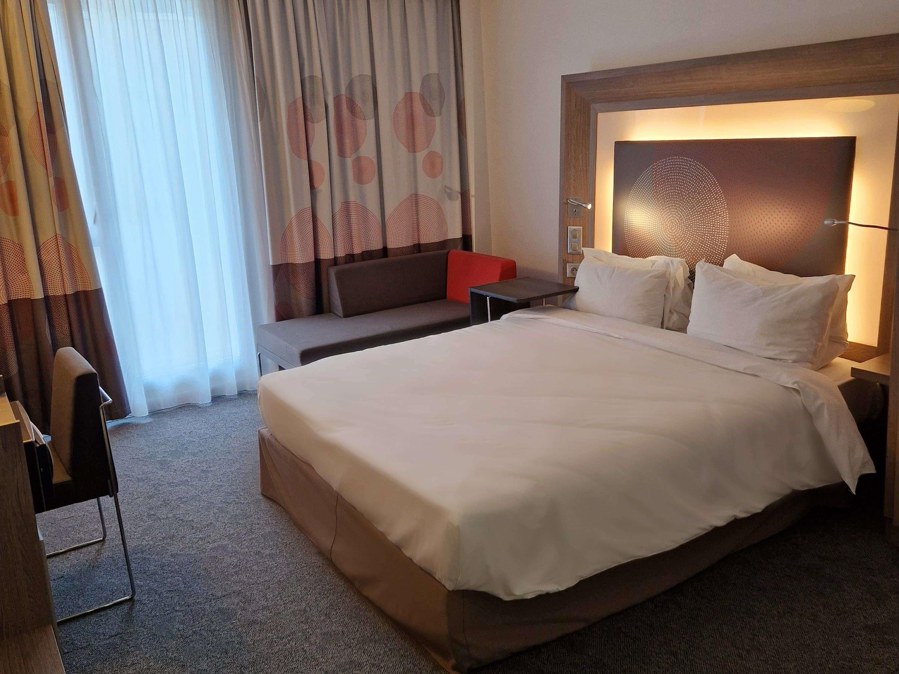 A hotel room with a large bed, a bedside table, a red chair, a sofa, and a wall-mounted lamp.