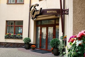 Entrada de hotel con un cartel que dice "Egel HOTEL", amplias puertas de vidrio y plantas con flores.