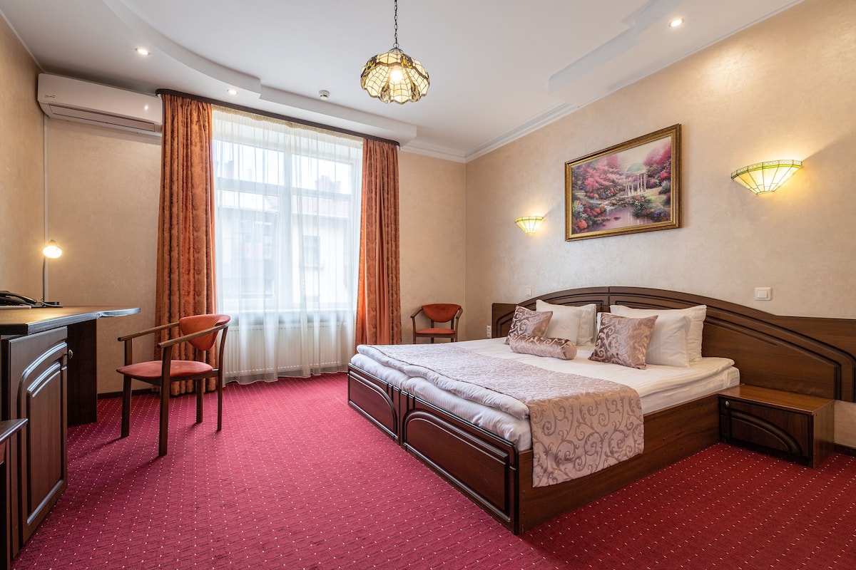 A hotel room with a bed, desk, chair, and a large window with curtains.