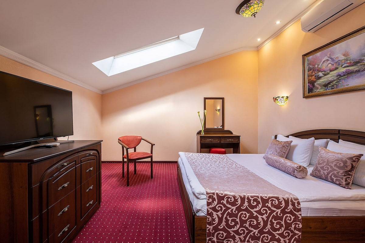 A hotel room with a bed, a desk, a chair, a TV, a dresser, a mirror, and a painting on the wall.