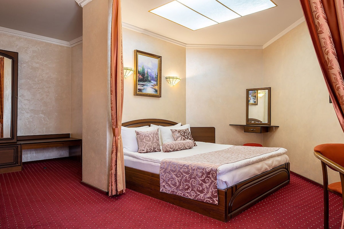A hotel room with a bed, a bedside table, a mirror, and a window with curtains.