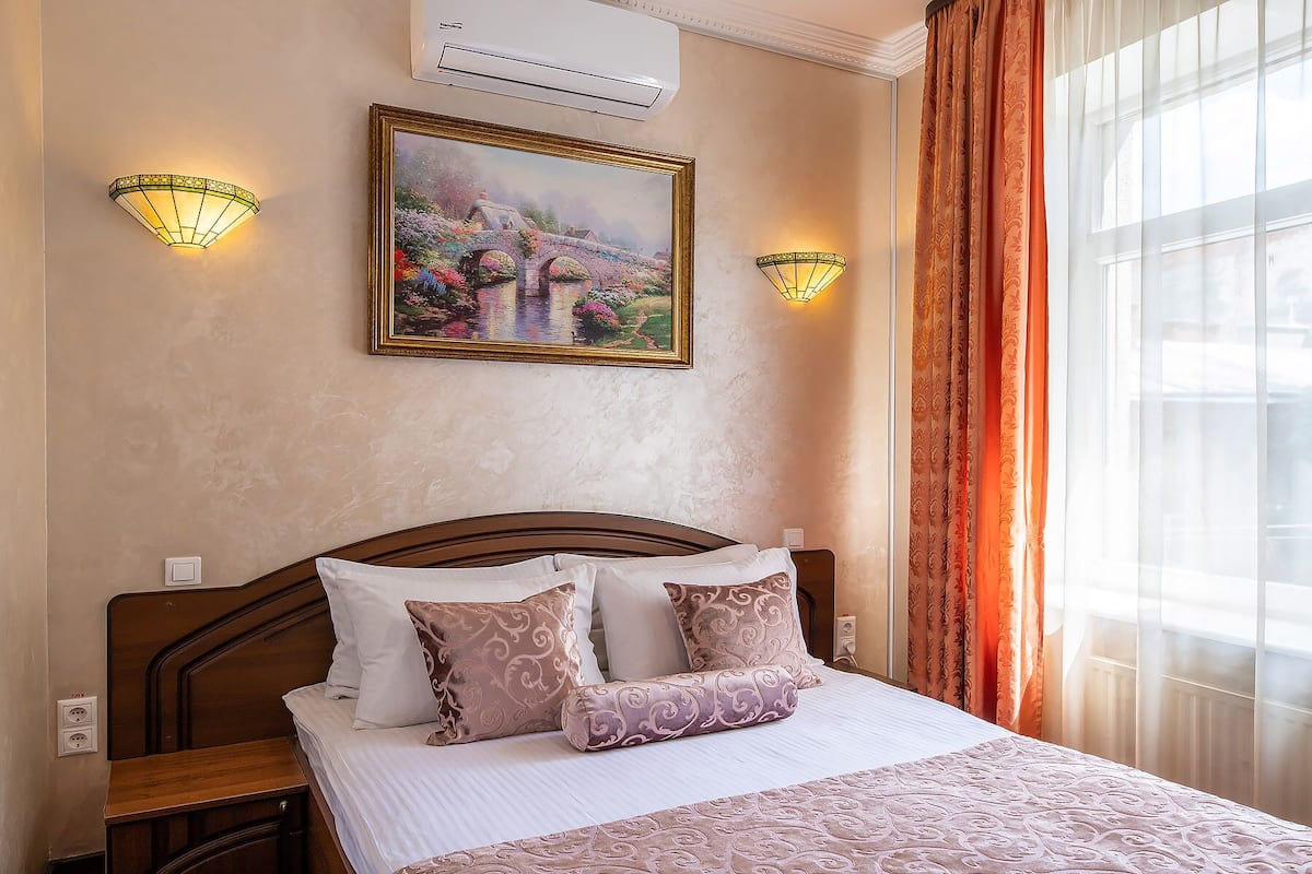 A hotel room with a bed, two wall-mounted lamps, an air conditioner, and a painting above the bed.