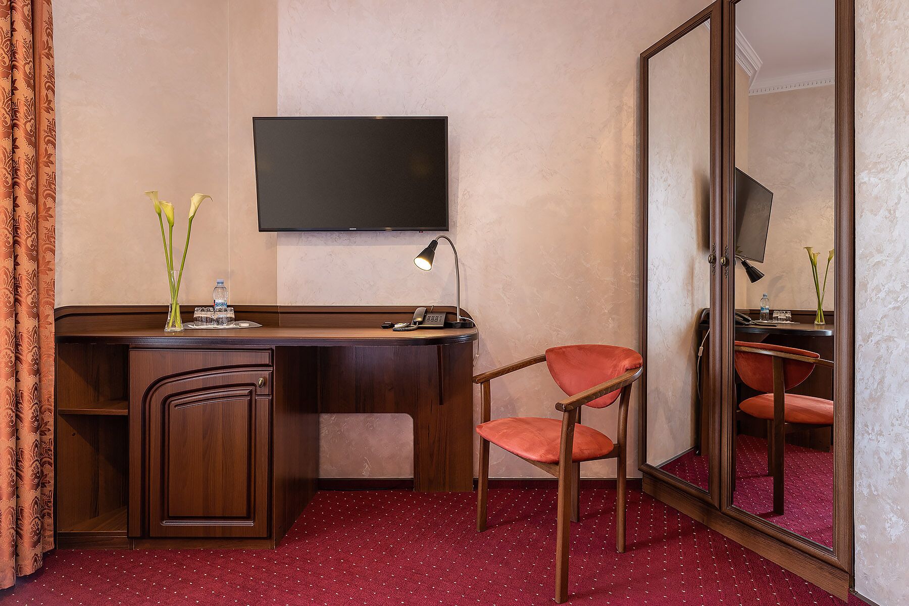 A hotel room with a wooden desk, a chair, a flat-screen TV mounted on the wall, a desk lamp, and a mirror.