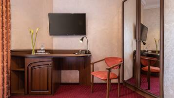 A hotel room with a wooden desk, a chair, a flat-screen TV mounted on the wall, a desk lamp, and a mirror.