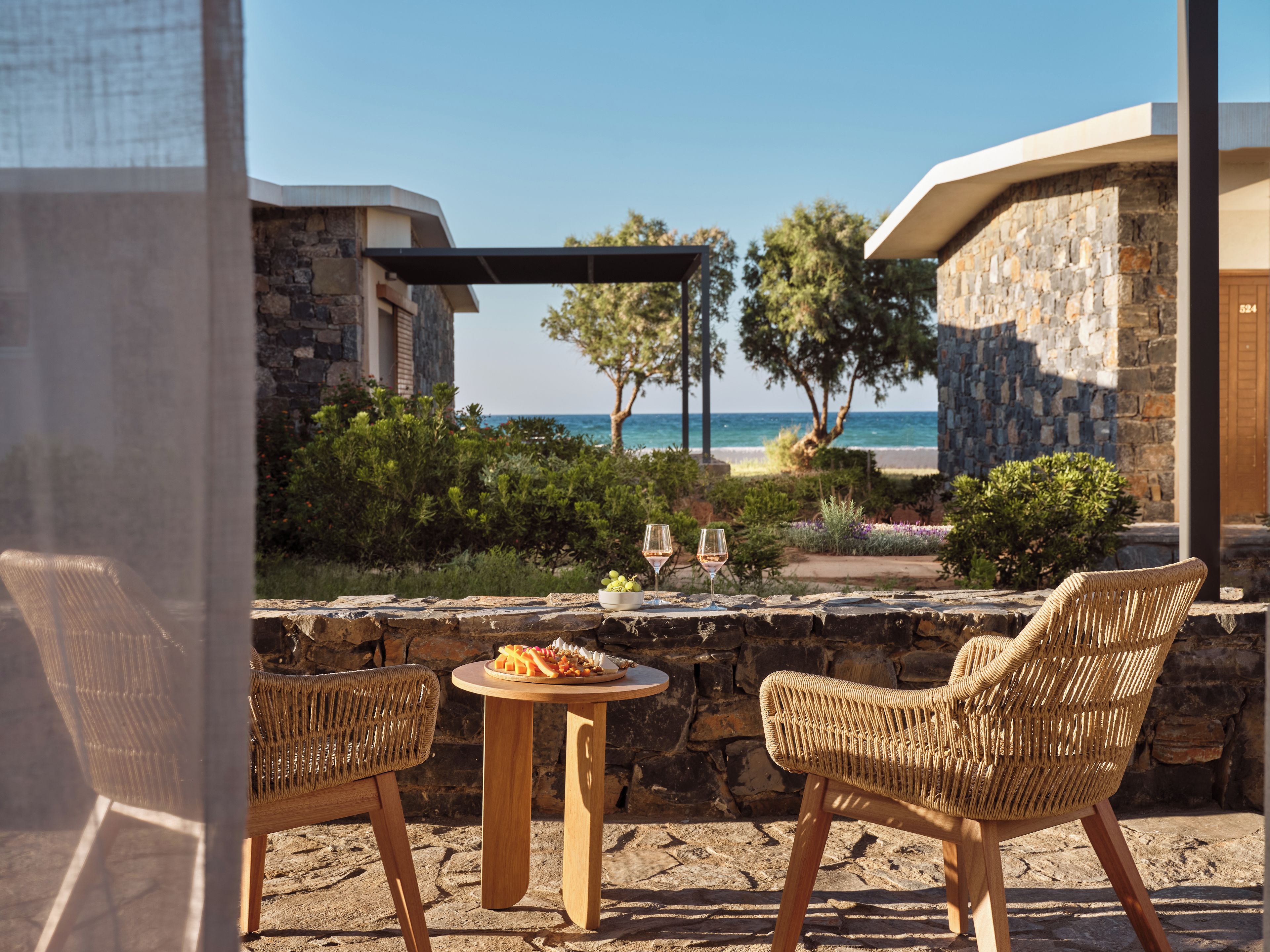 Eine Terrasse mit Korbstühlen und einem Tisch, mit Blick auf einen Strand und ein Steinhaus.