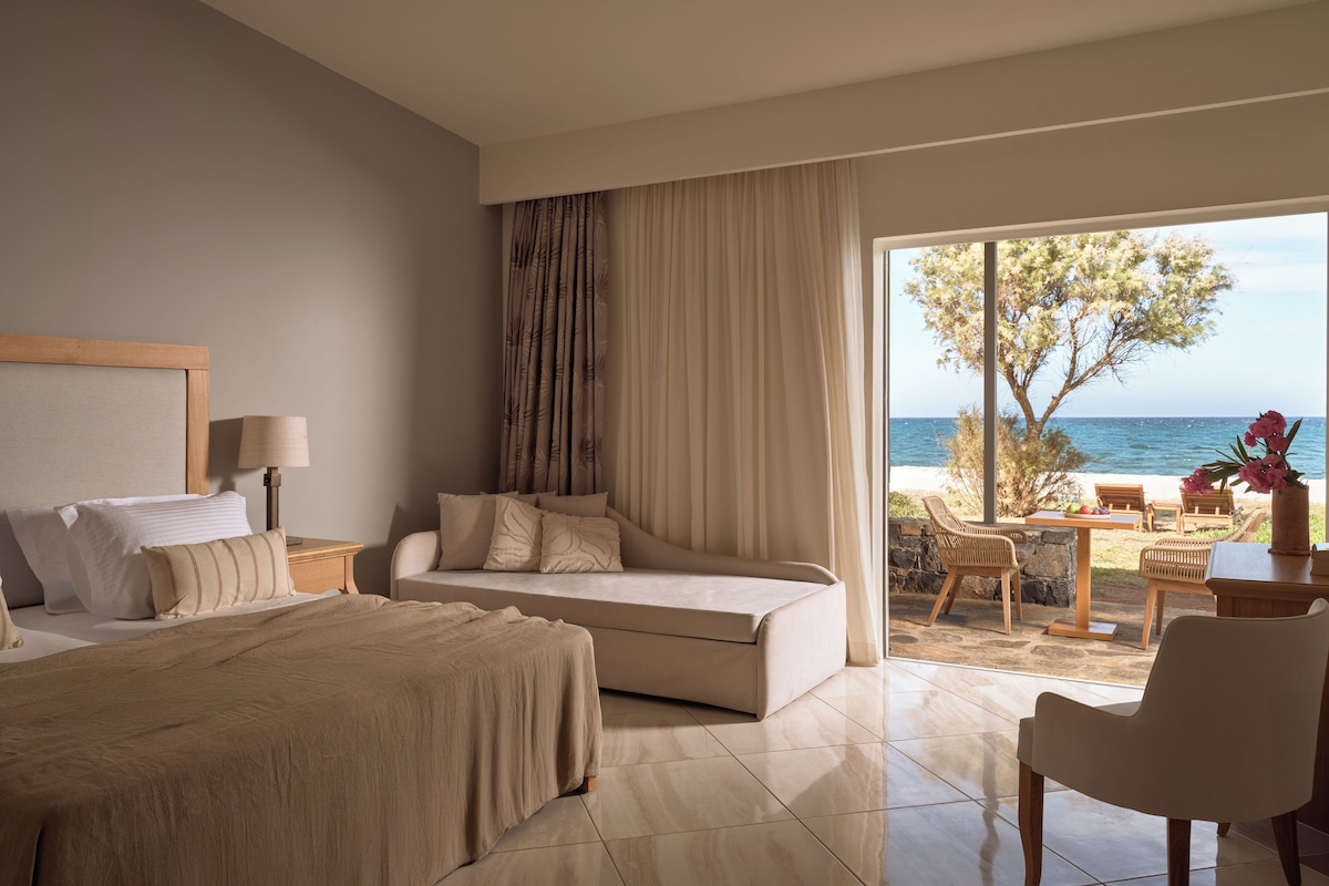A hotel room with a bed, a sofa, a dining area with chairs, and a view of the sea.