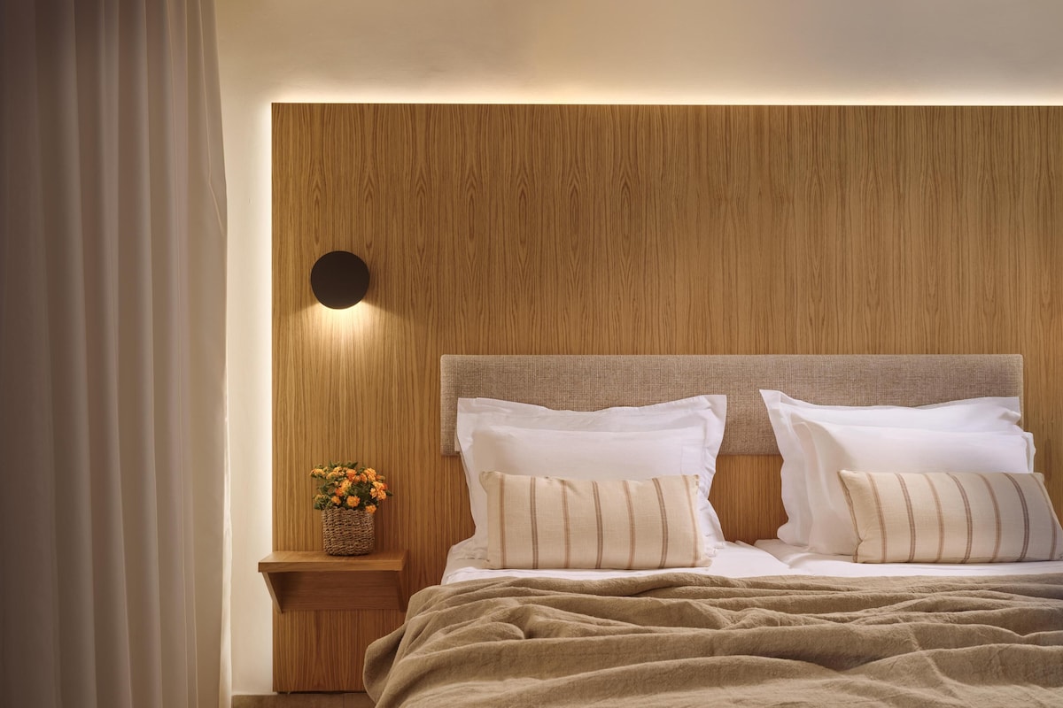 A neatly made bed with a wooden headboard, a bedside table with a vase of flowers, and a wall with a circular light fixture.