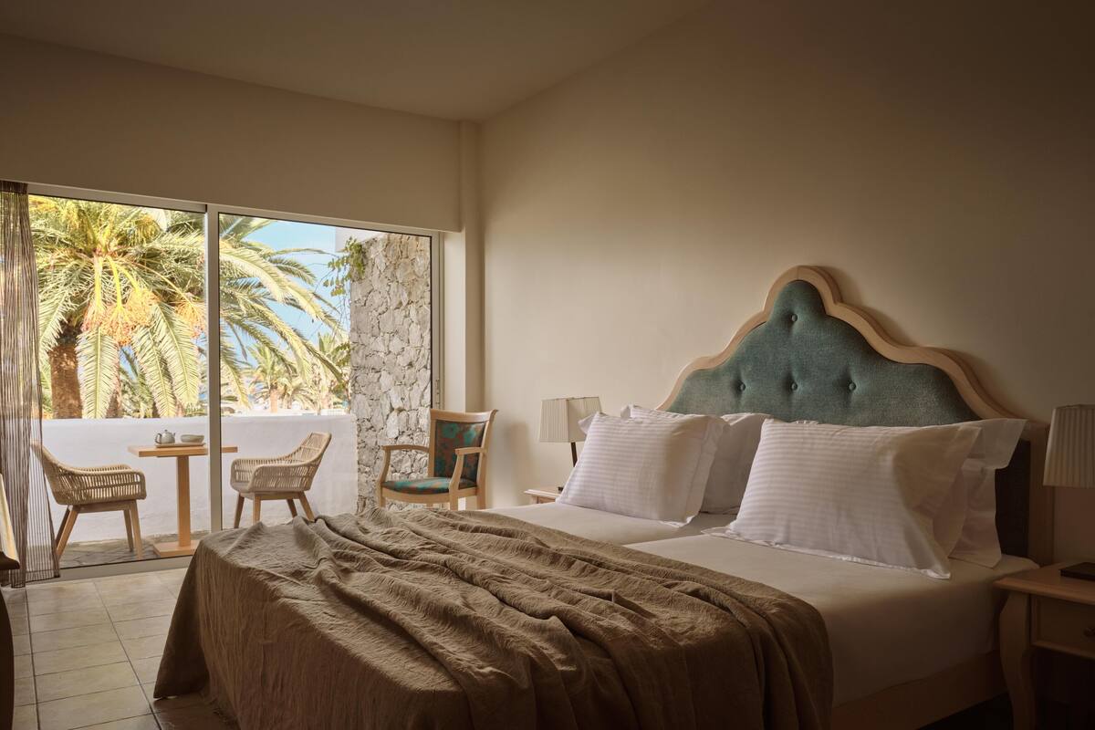 A hotel room with a large bed, a view of palm trees, and a balcony with wicker furniture.