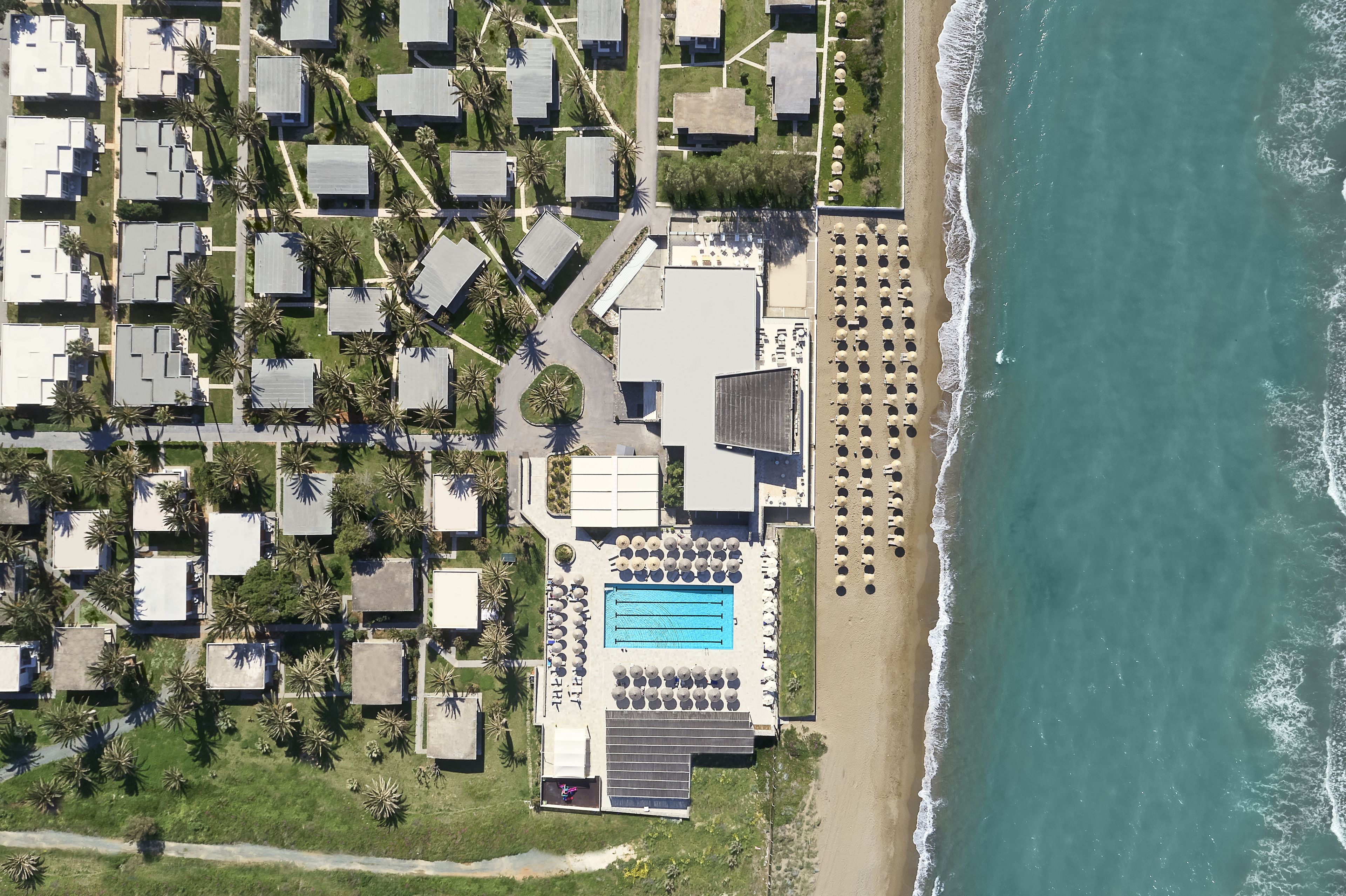 Eine Vogelperspektive zeigt einen Küstenort mit mehreren bungalowsartigen Unterkünften, einem zentral gelegenen Schwimmbad und einem Strandbereich.