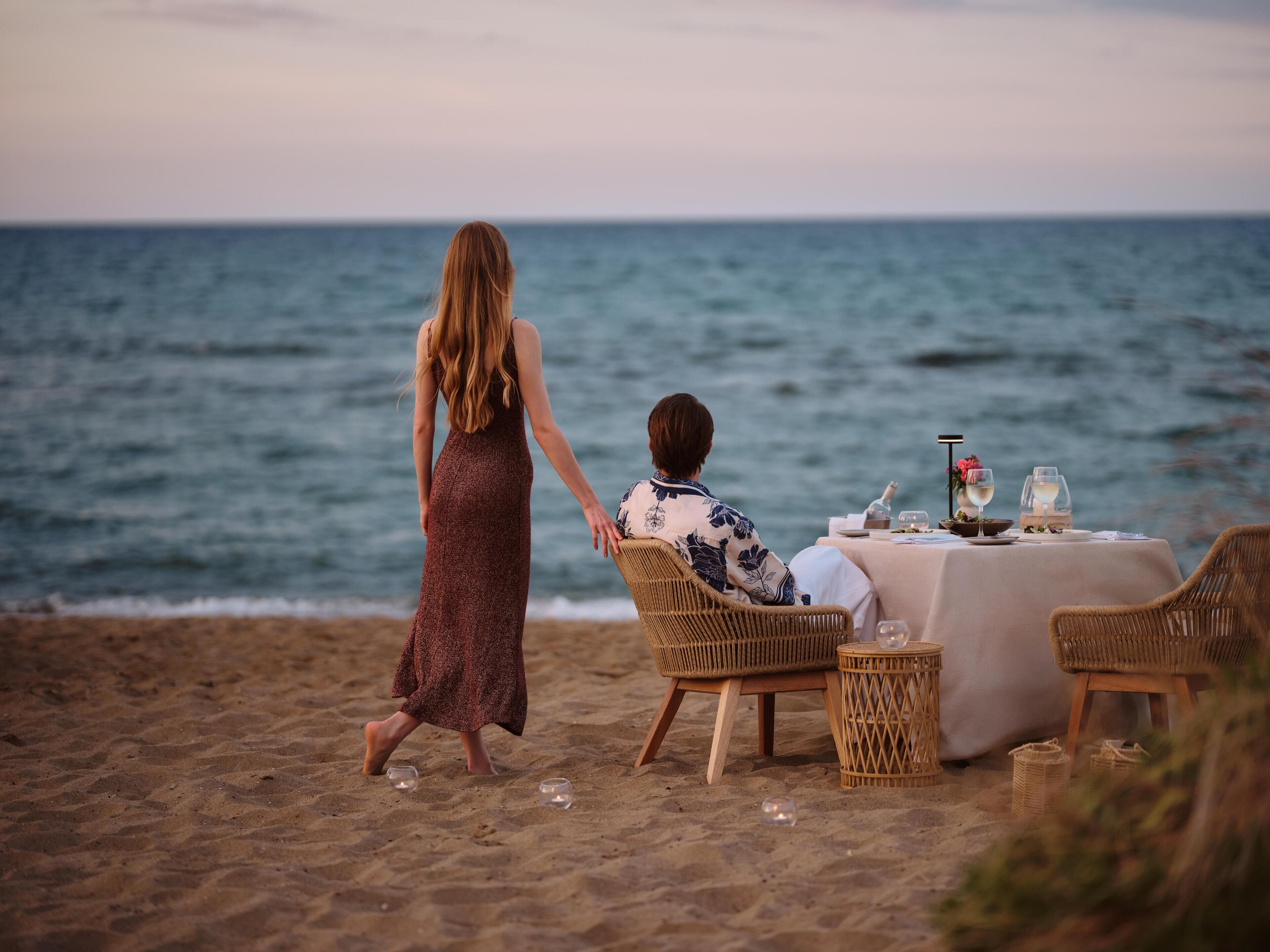 A couple walking on a beach with a table set for two, overlooking the sea.