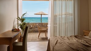 Una camera d'albergo con un letto, una scrivania in legno, una sedia, un tavolo da pranzo con della frutta e vista sull'oceano attraverso una finestra con tende.
