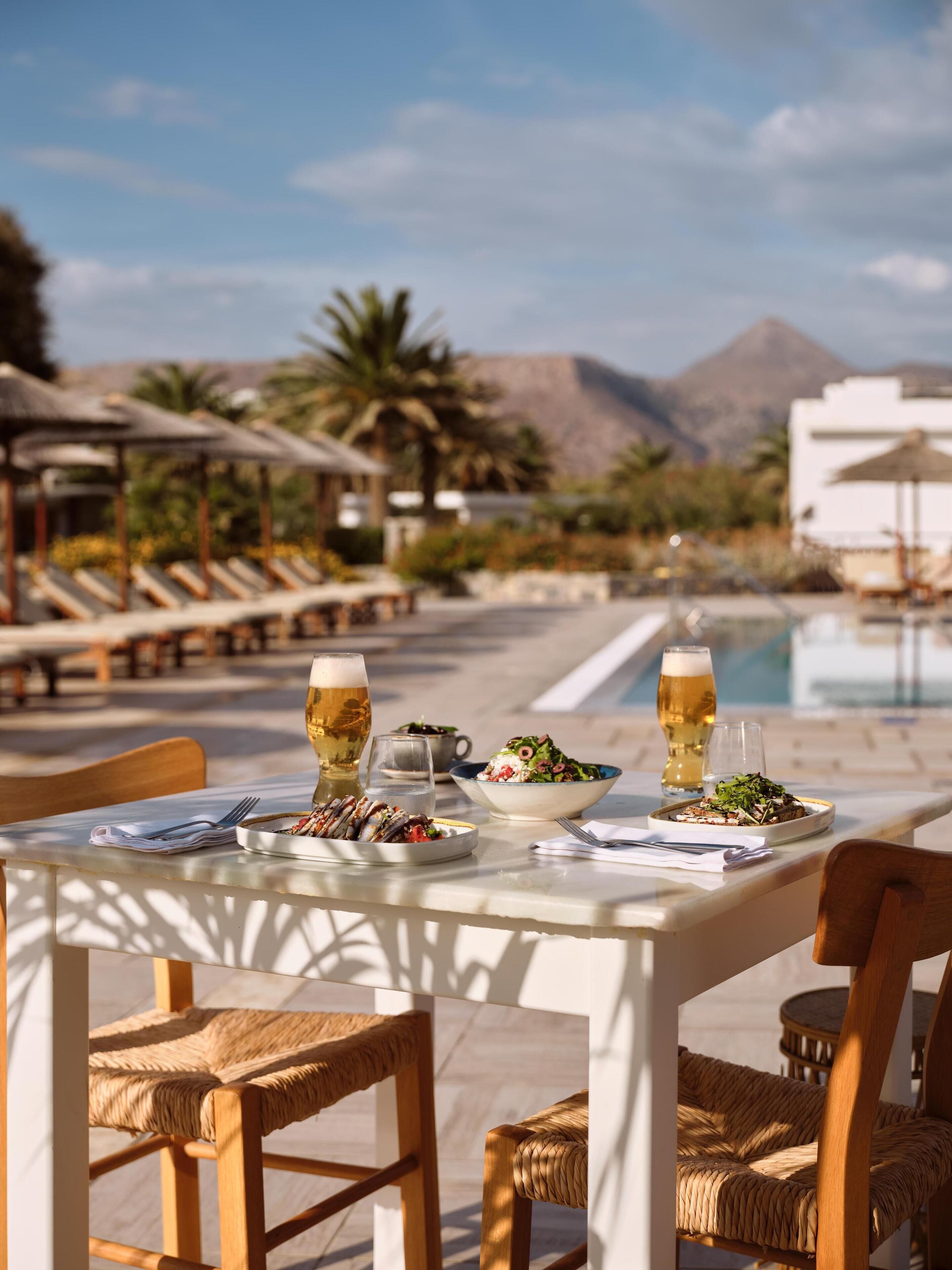 A dining table with plates of food and drinks, surrounded by wicker chairs, set against a poolside view with mountains in the background.