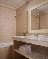 A bathroom with beige tiles, a white sink, a mirror, a shower, and a shelf with folded towels.