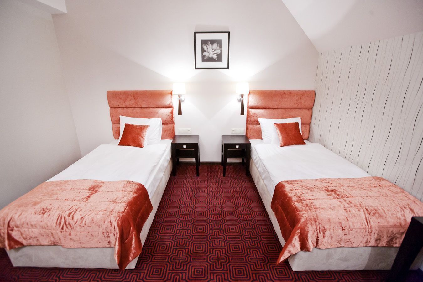 Habitación de hotel con dos camas, cada una con cabecero rojo, ropa de cama blanca y alfombra roja estampada. Hay dos mesitas de noche con lámparas, y un cuadro enmarcado en la pared.