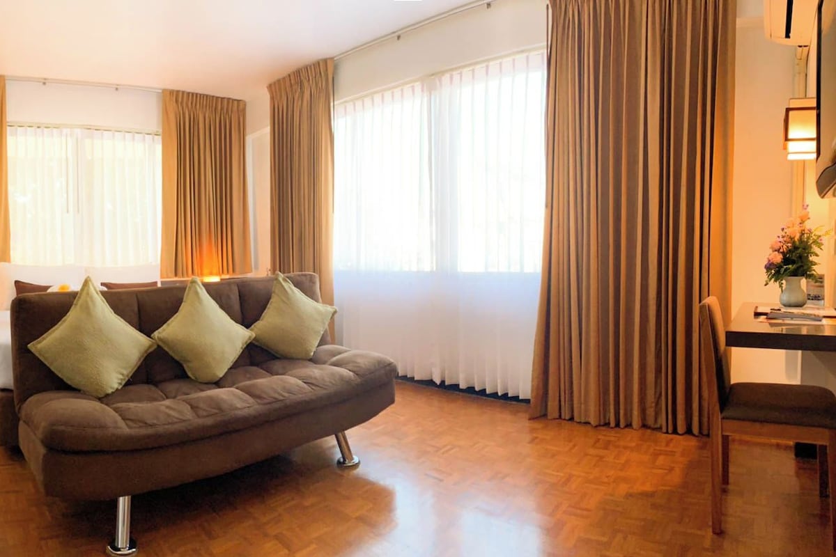 A modern living room with a brown sofa, a small table, and a chair. There are curtains on the windows, and a lamp is on the table.