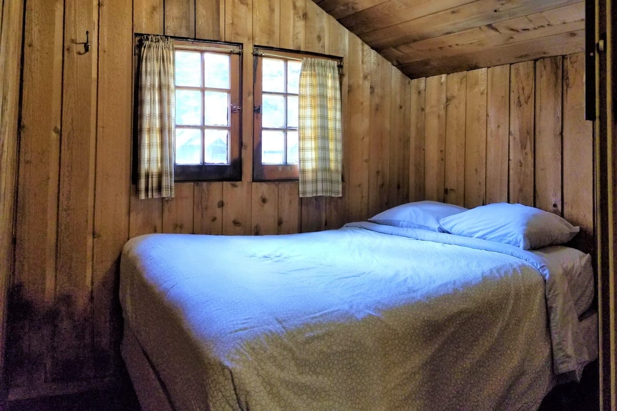 A wooden cabin room with a bed, two windows with curtains, and a light fixture on the ceiling.