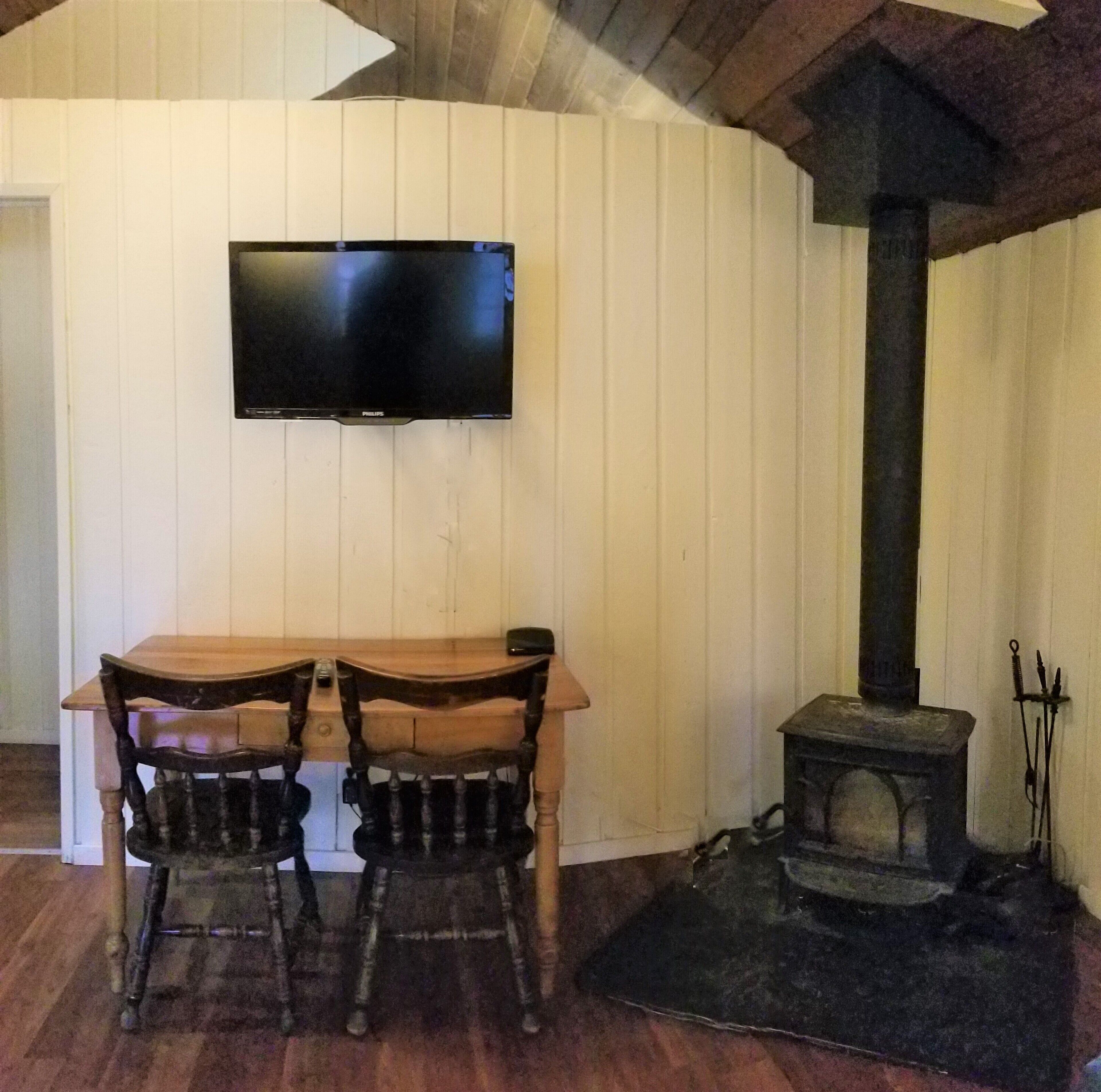 A room with a wooden table and chairs, a television mounted on the wall, and a wood-burning stove.