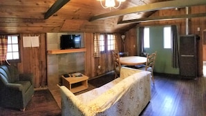A wooden cabin interior with a sofa, a television on a shelf, a dining table, and a refrigerator.