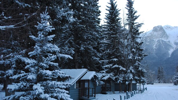 A snow-covered lodge with multiple cabins, surrounded by tall evergreen trees and mountains in the background.