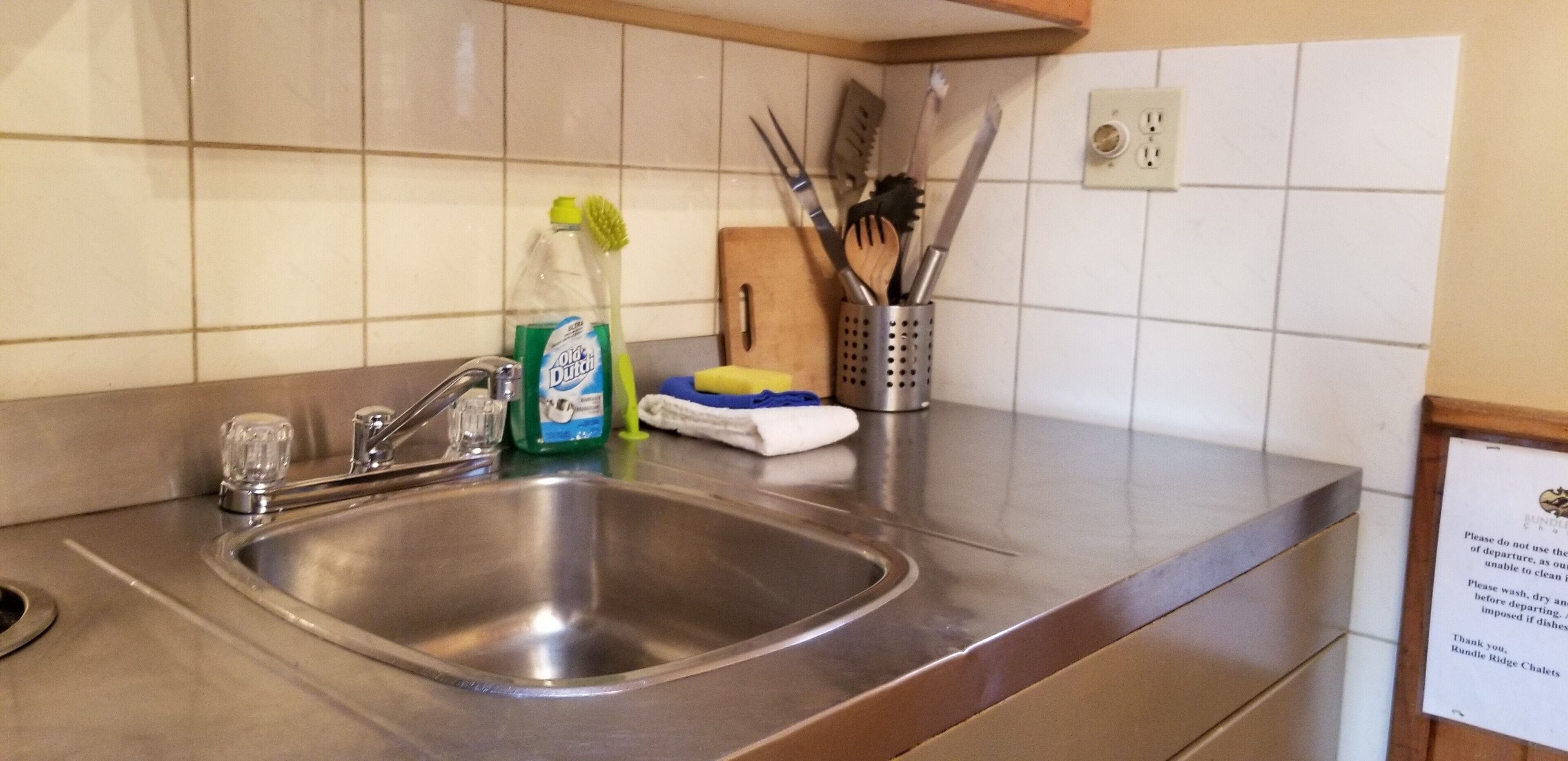 Un lavamanos de acero inoxidable con un grifo, una botella de detergente, una esponja y un recipiente con utensilios.
