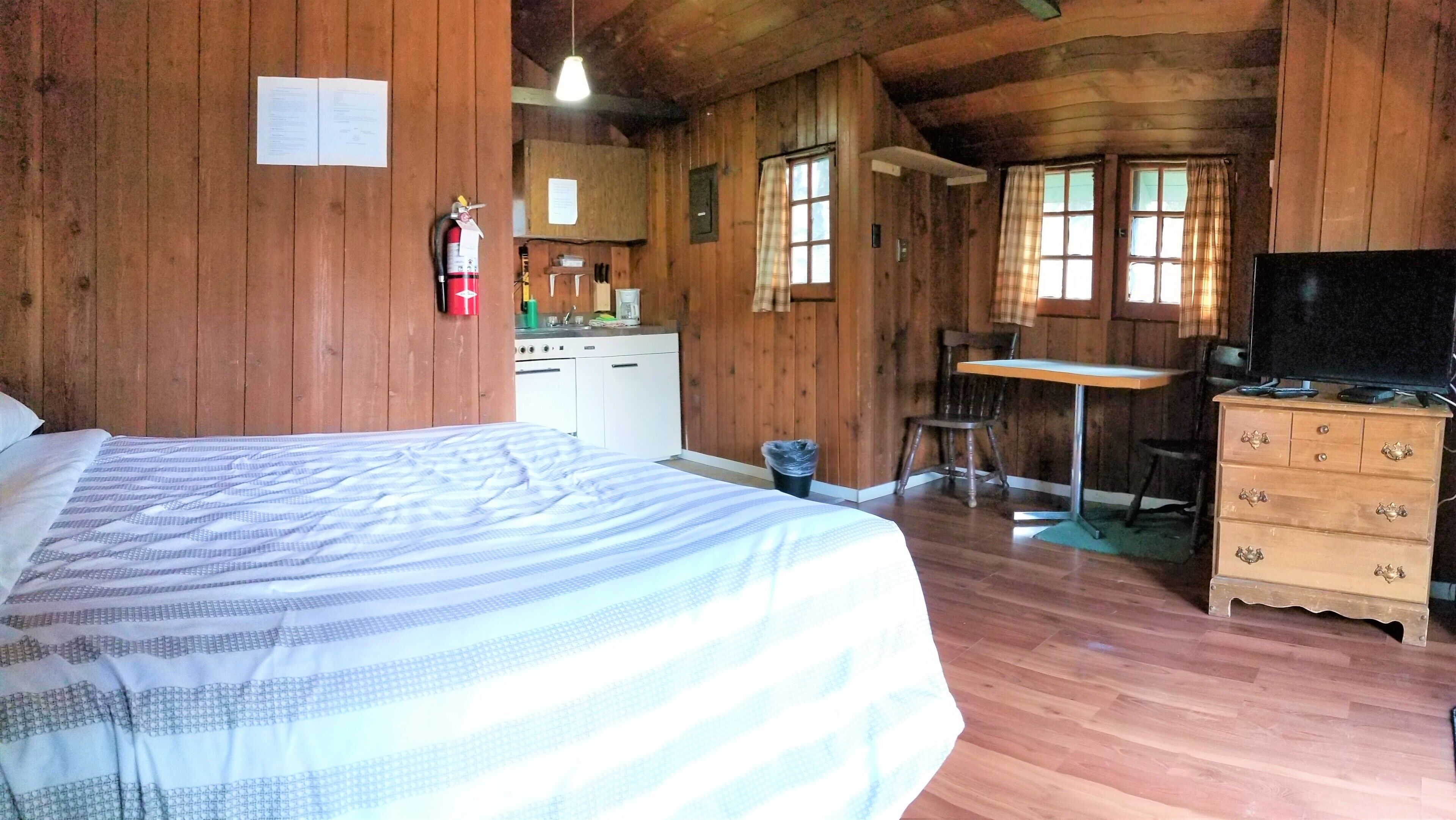 Ein Zimmer in einer Holzhütte mit einem Bett, einem Schreibtisch, einem Sessel, einem Schminktisch und einem Fernseher.