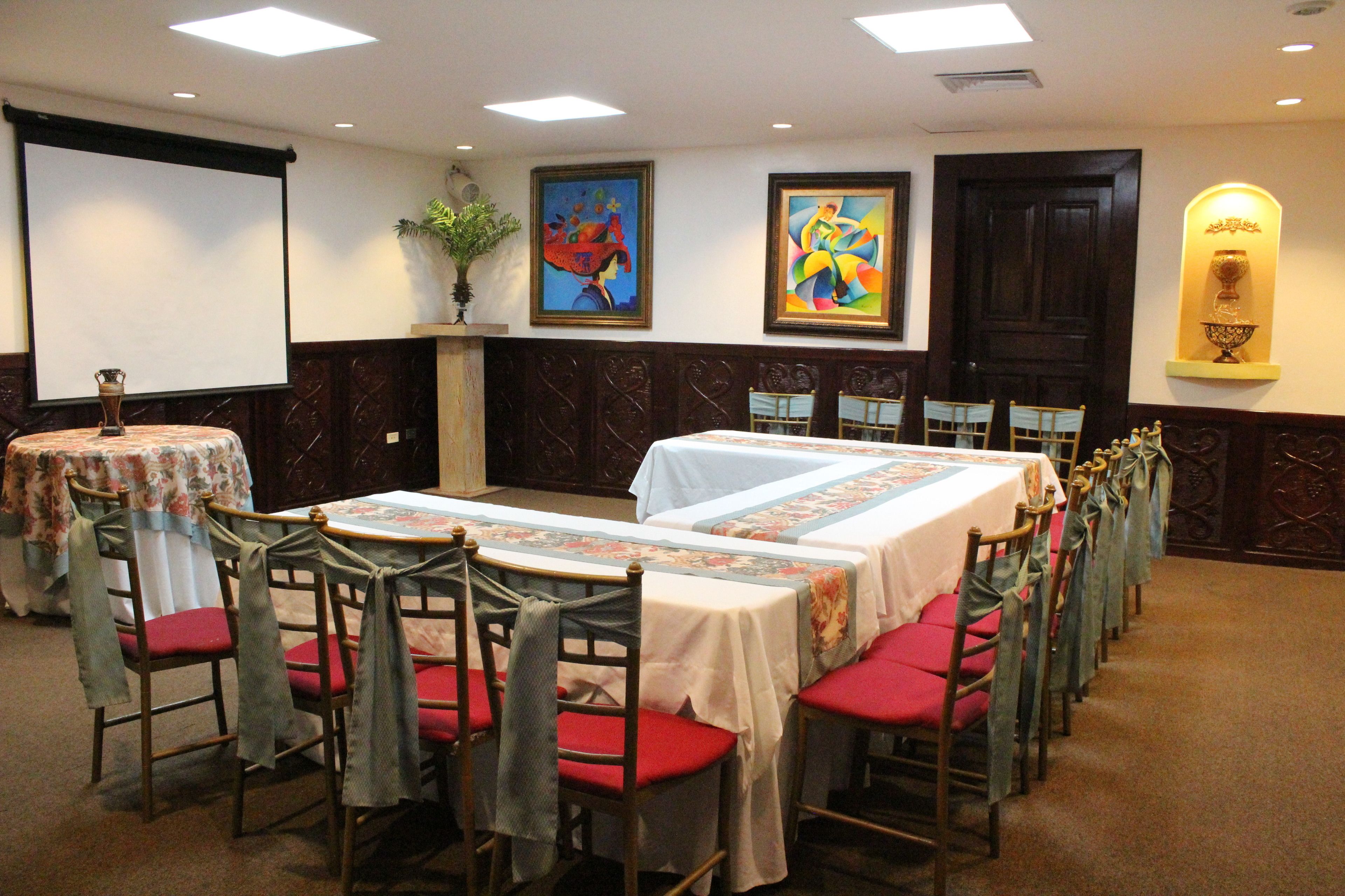 Un salón de conferencias con mesas y sillas, una pantalla de proyector y obras de arte en las paredes.
