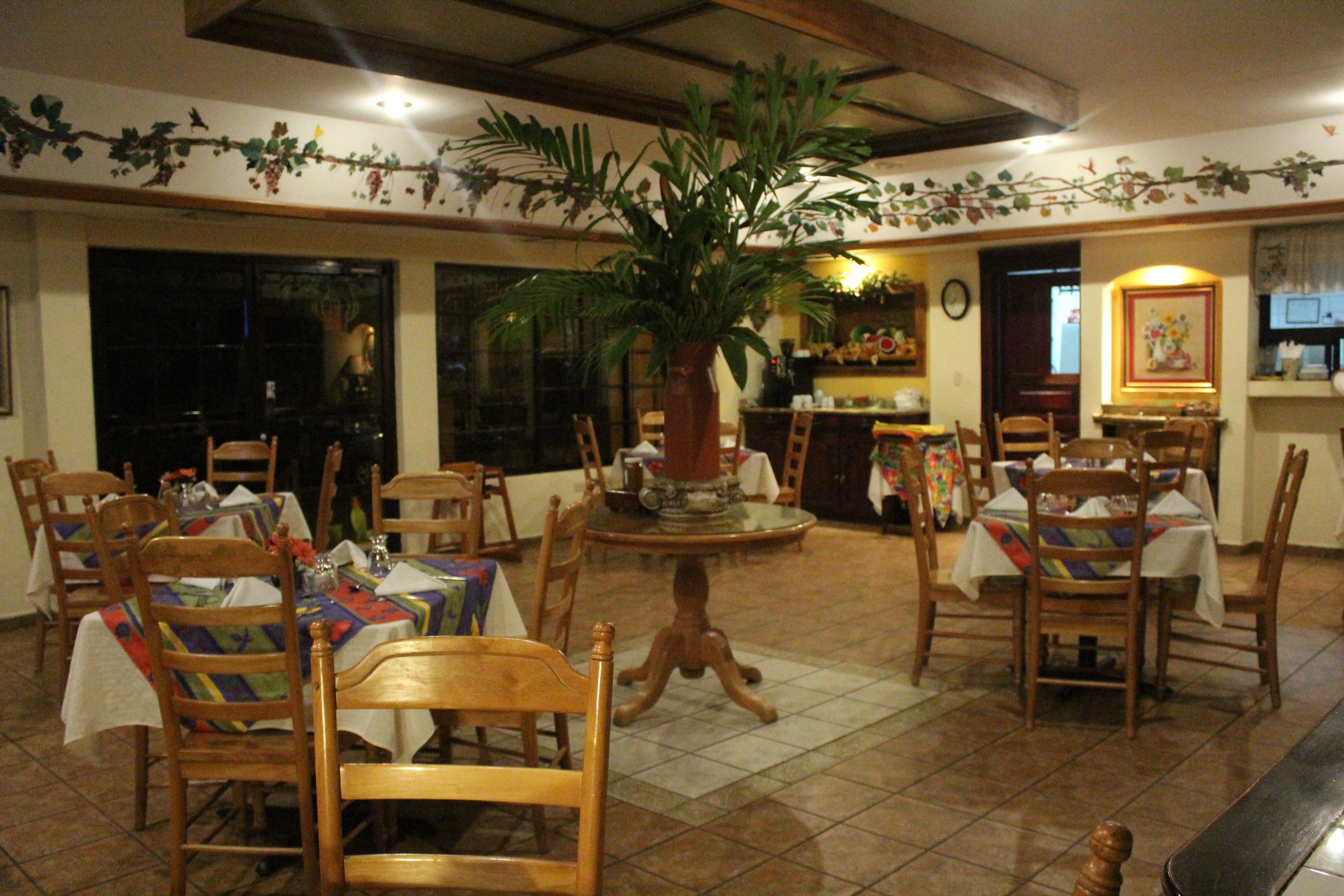 A dining area with wooden chairs and tables, a large potted plant, and a tiled floor.