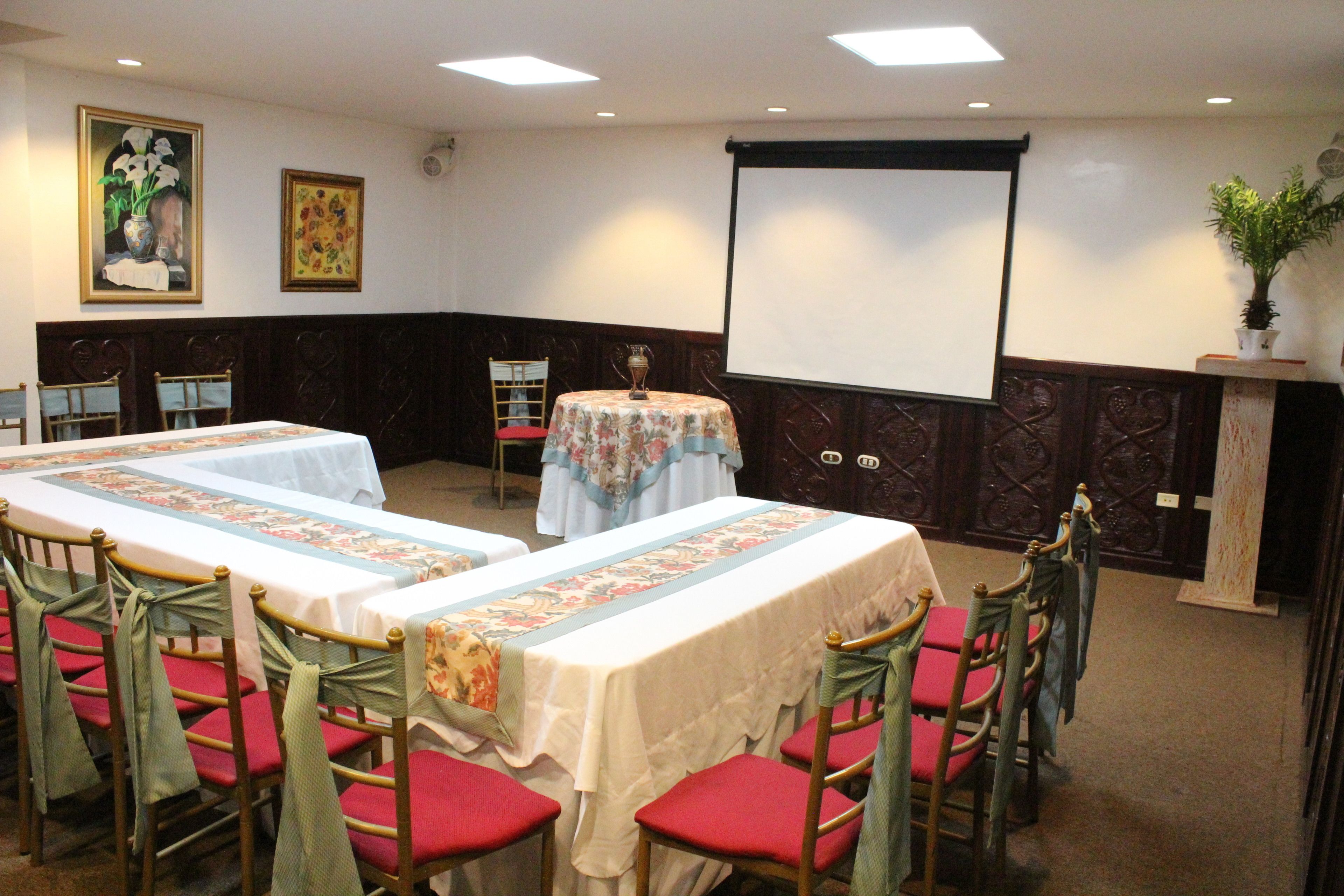 A conference room with two long tables, red chairs, and a projector screen.
