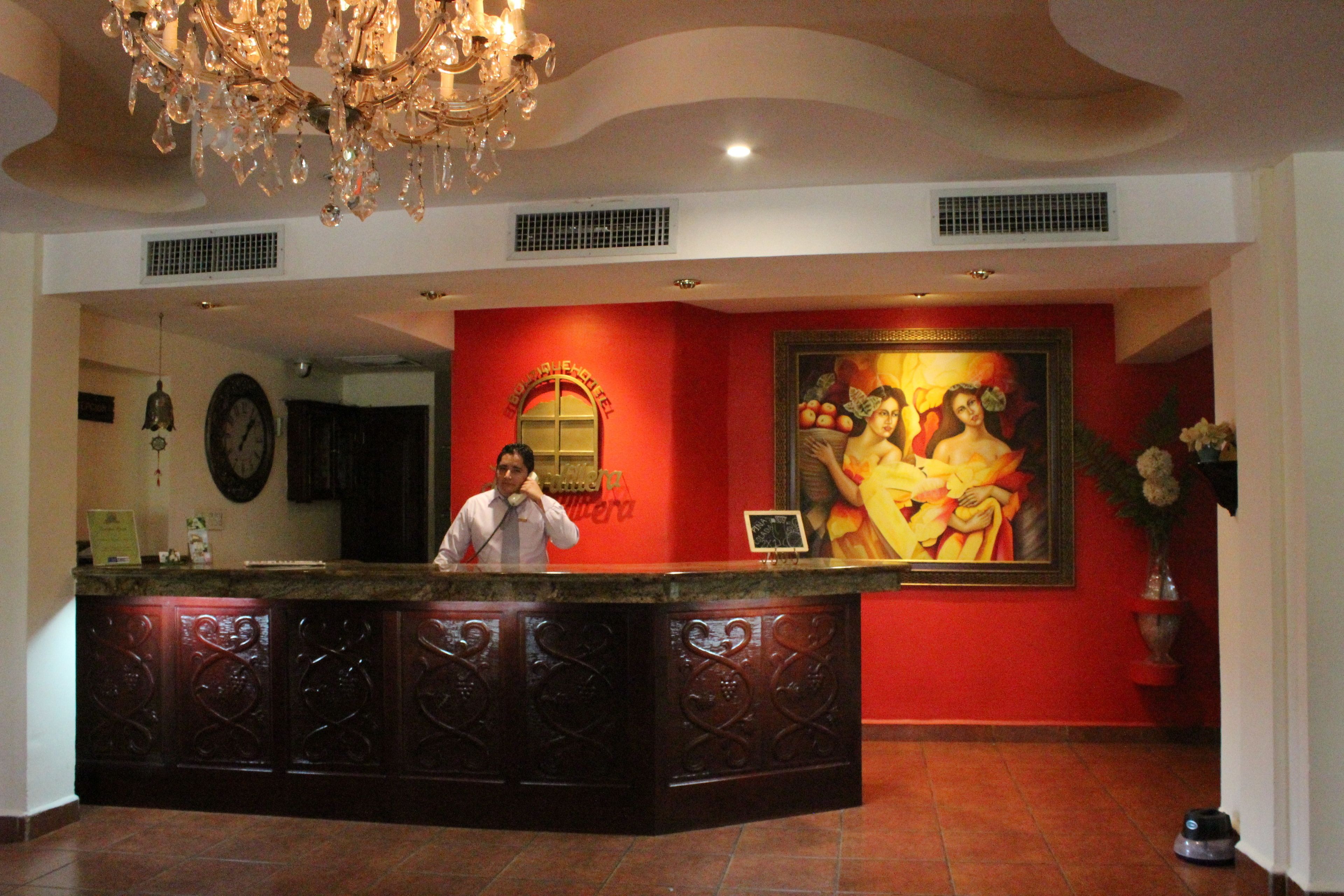 A reception desk with a man standing behind it, a large painting on the wall, and a chandelier above.