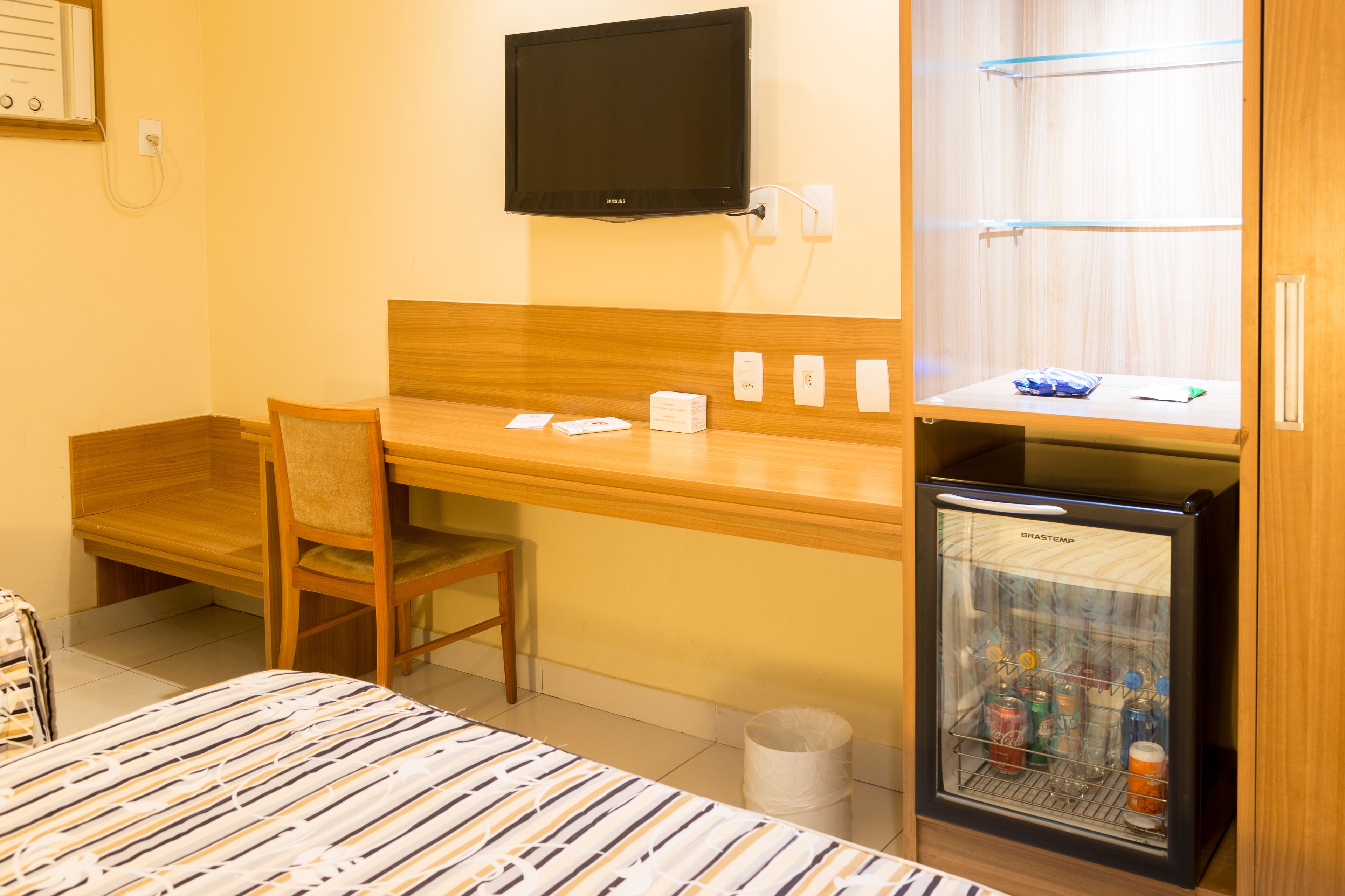 A hotel room with a bed, a desk with a chair, a television, a minibar, and a trash bin.