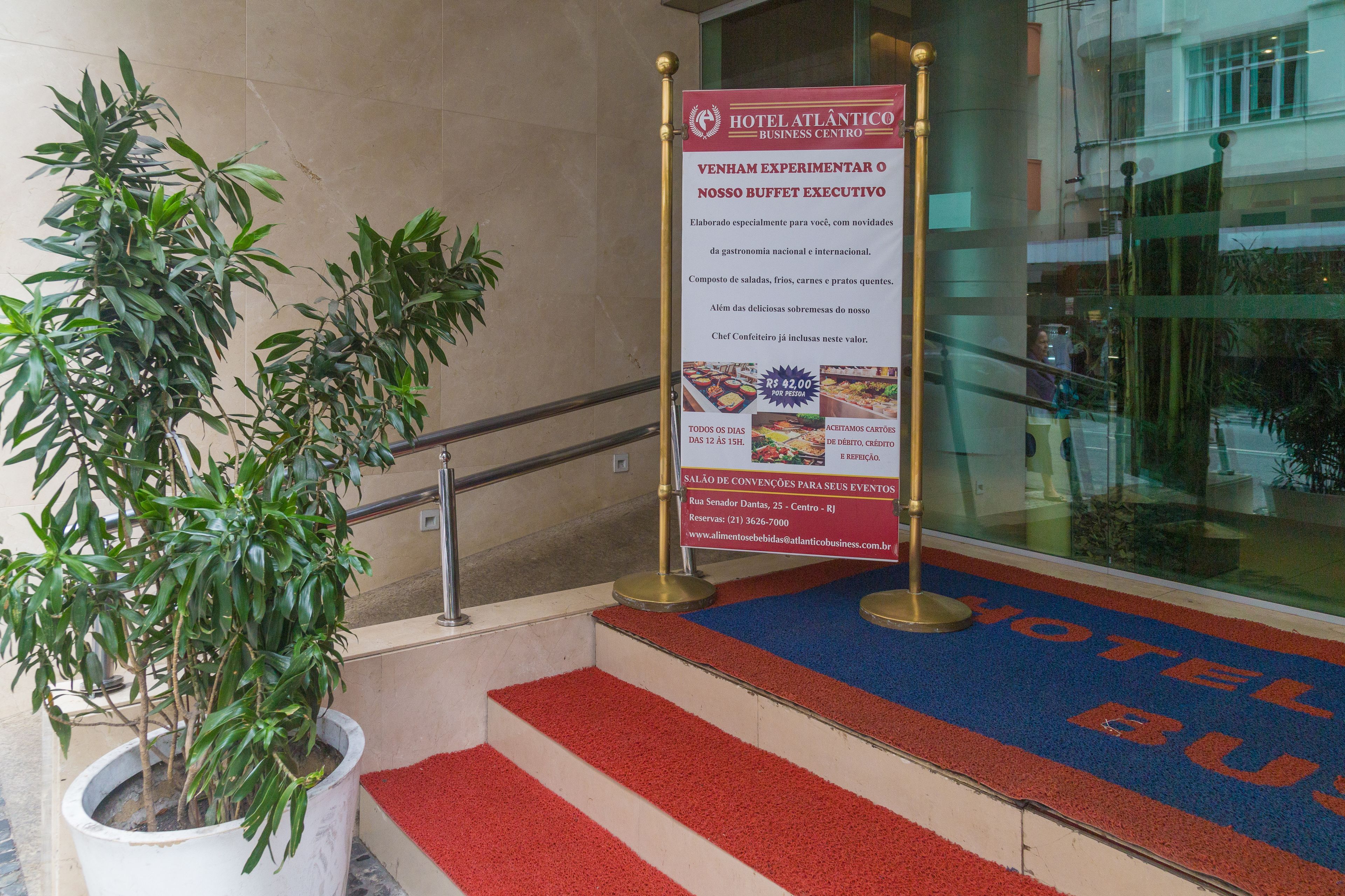 Entrada de un hotel con alfombra roja que conduce al vestíbulo, un cartel anunciando el bufé ejecutivo del hotel y una planta en maceta junto a las escaleras.