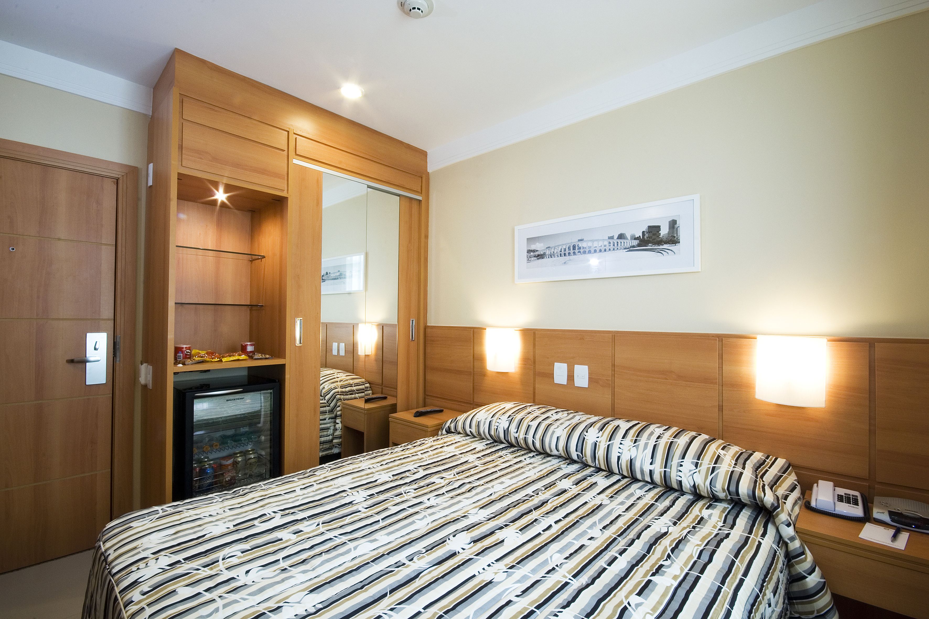 Een hotelkamer met een bed, een koelkast, een nachtkastje met een lamp en een lijst aan de muur.
