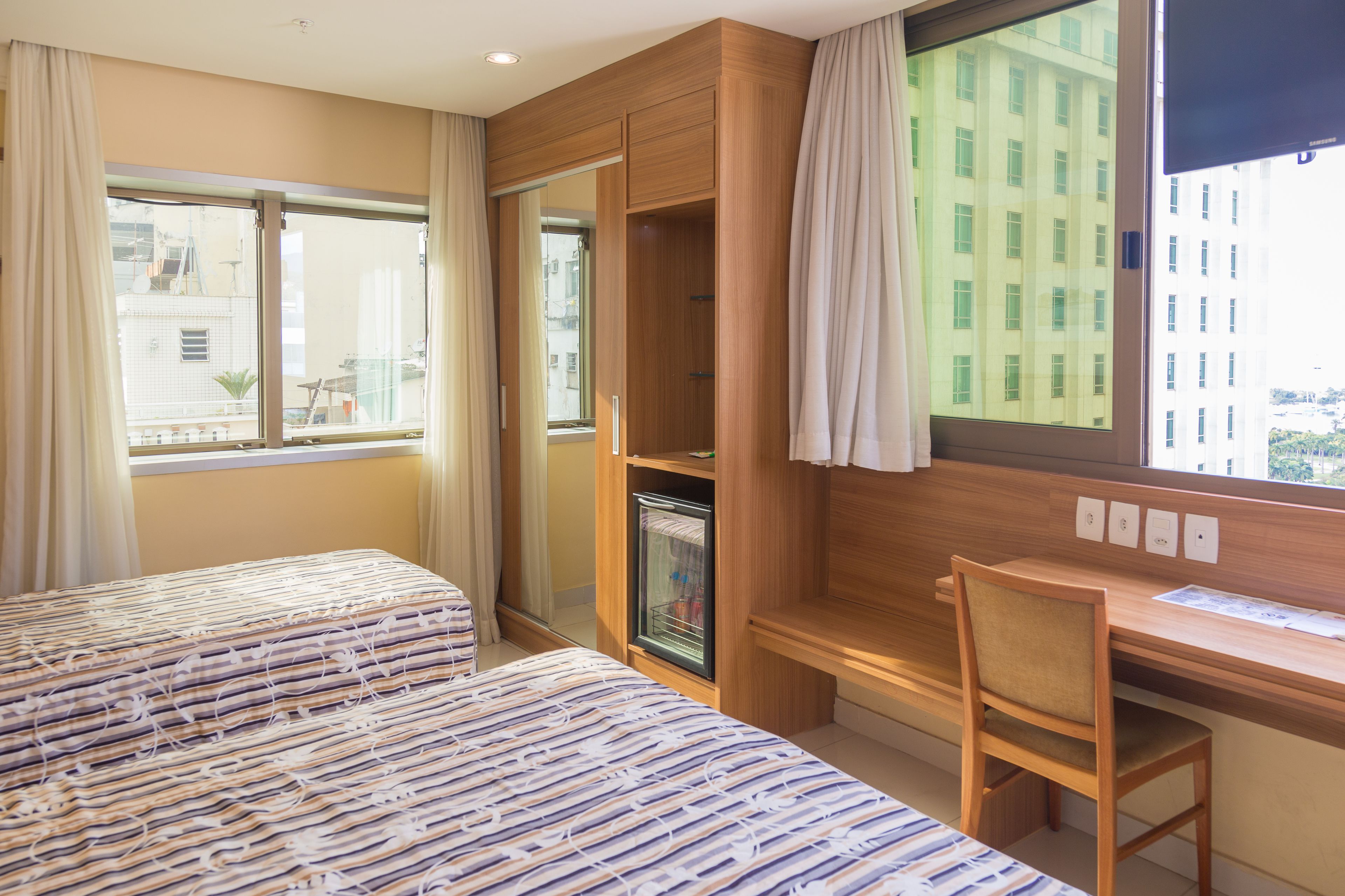 Quarto de hotel com duas camas, uma mesa e vista para um edifício através da janela.