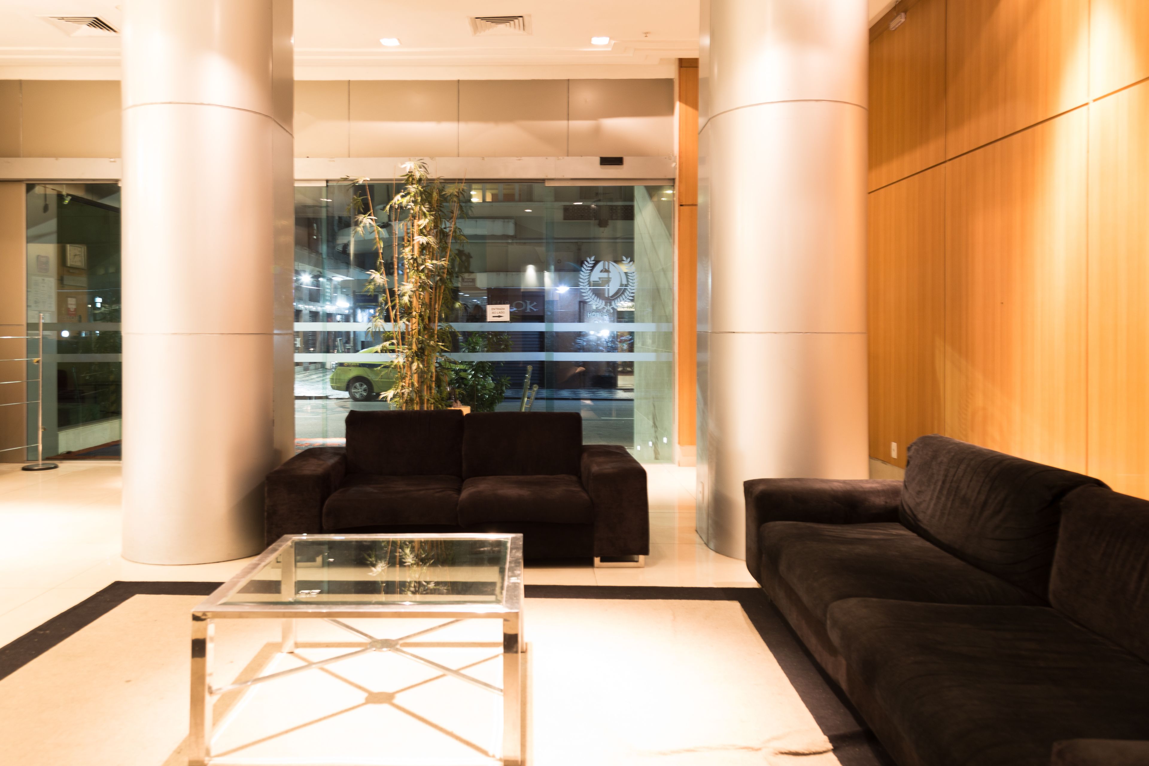 A modern lobby with a glass coffee table, two dark sofas, and a tall plant.