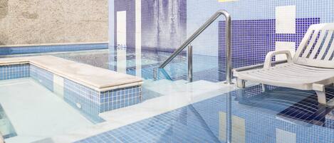 A swimming pool with a tiled floor and walls, a metal ladder, and a white lounge chair.