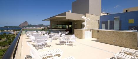 Una terraza al aire libre con sillones blancos y mesas, un edificio moderno con fachada azul y vistas a la montaña y al océano.