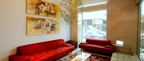 A modern living room with red sofas, a glass coffee table, abstract paintings, and a large window with a view of the street.