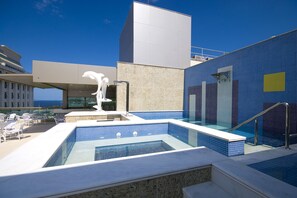 Een dakzwembad met helderblauwe tegels, een witte sculptuur en een modern gebouw met een glazen gevel.