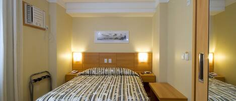 Una habitación de hotel con cama, mesitas de noche, escritorio, silla, espejo y un cuadro en la pared.