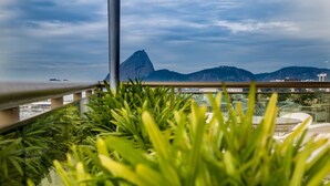 Un balcon agrémenté de plantes luxuriantes, doté d’une rambarde en métal, et offrant une vue sur une montagne et le paysage urbain en arrière-plan.