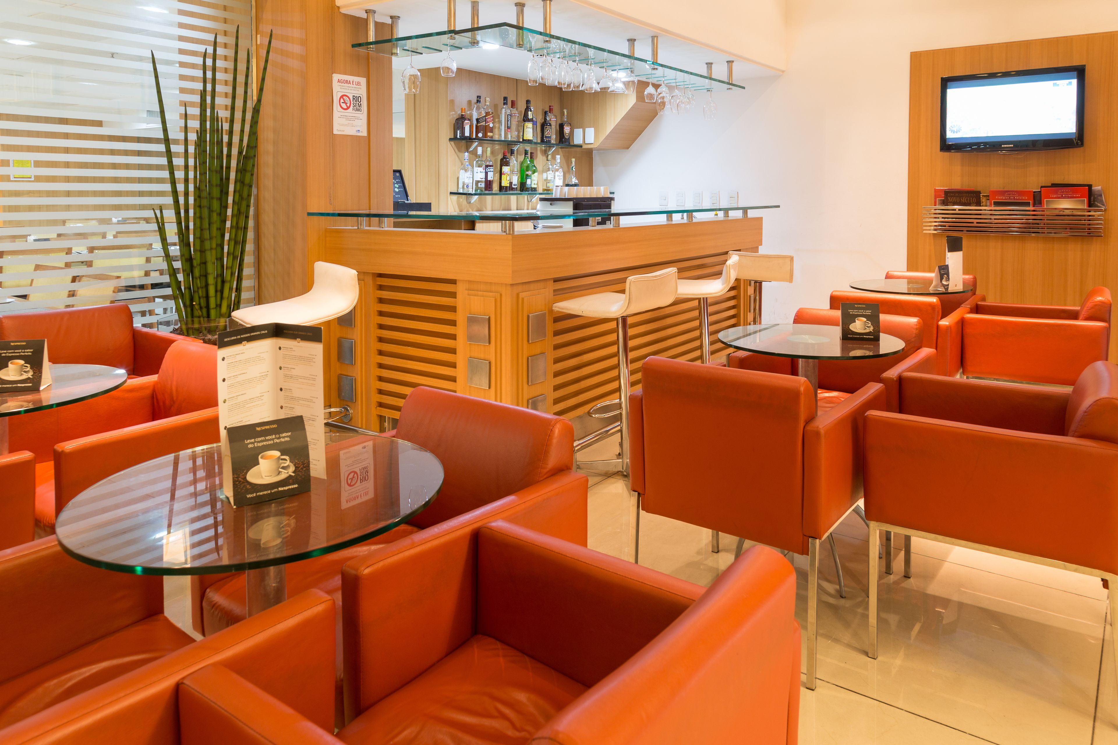 A modern bar area with glass tables and orange chairs, a bar counter with various bottles, and a TV mounted on the wall.