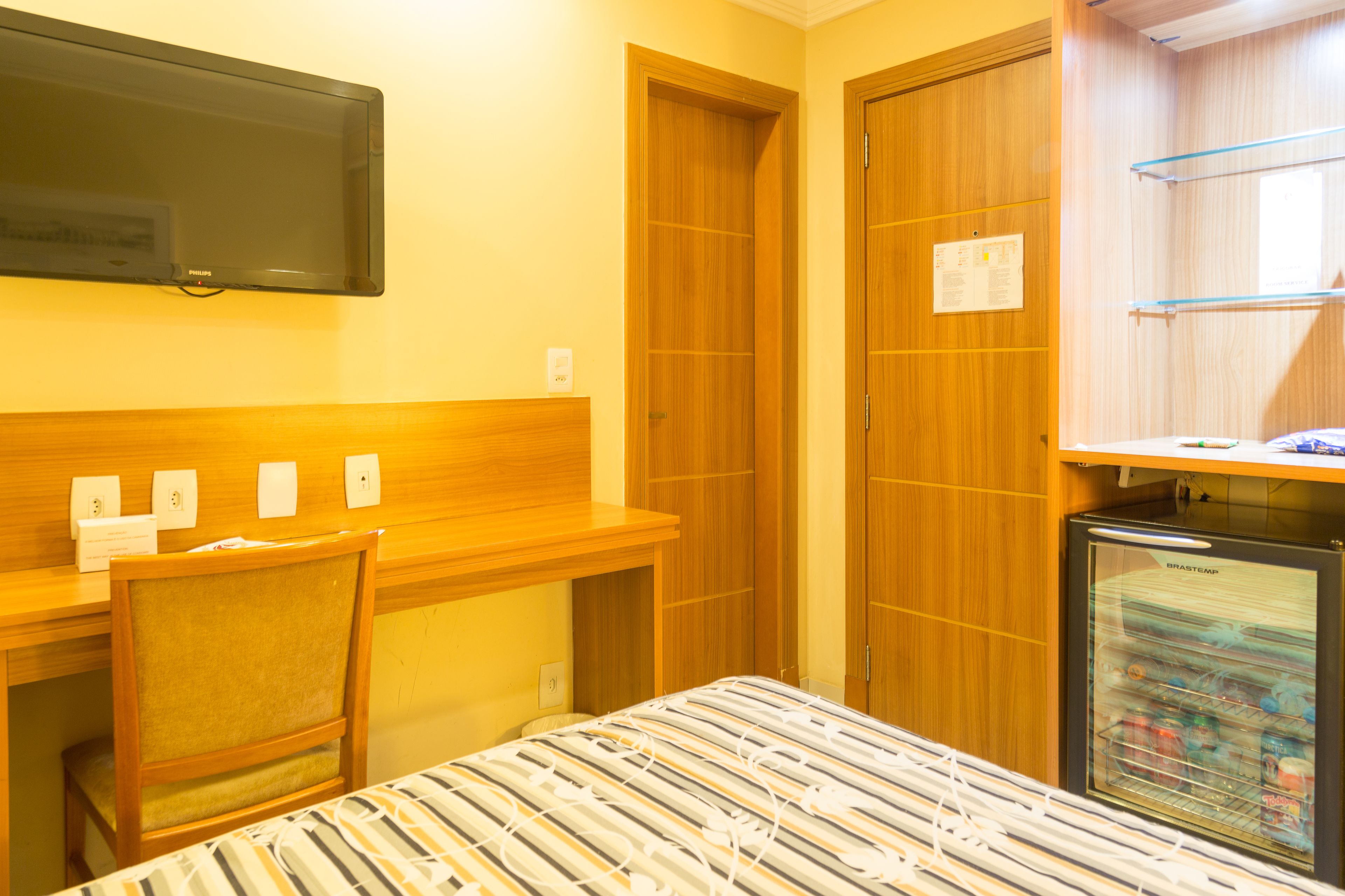 A hotel room with a bed, a desk, a chair, a television, a refrigerator, and wooden doors.