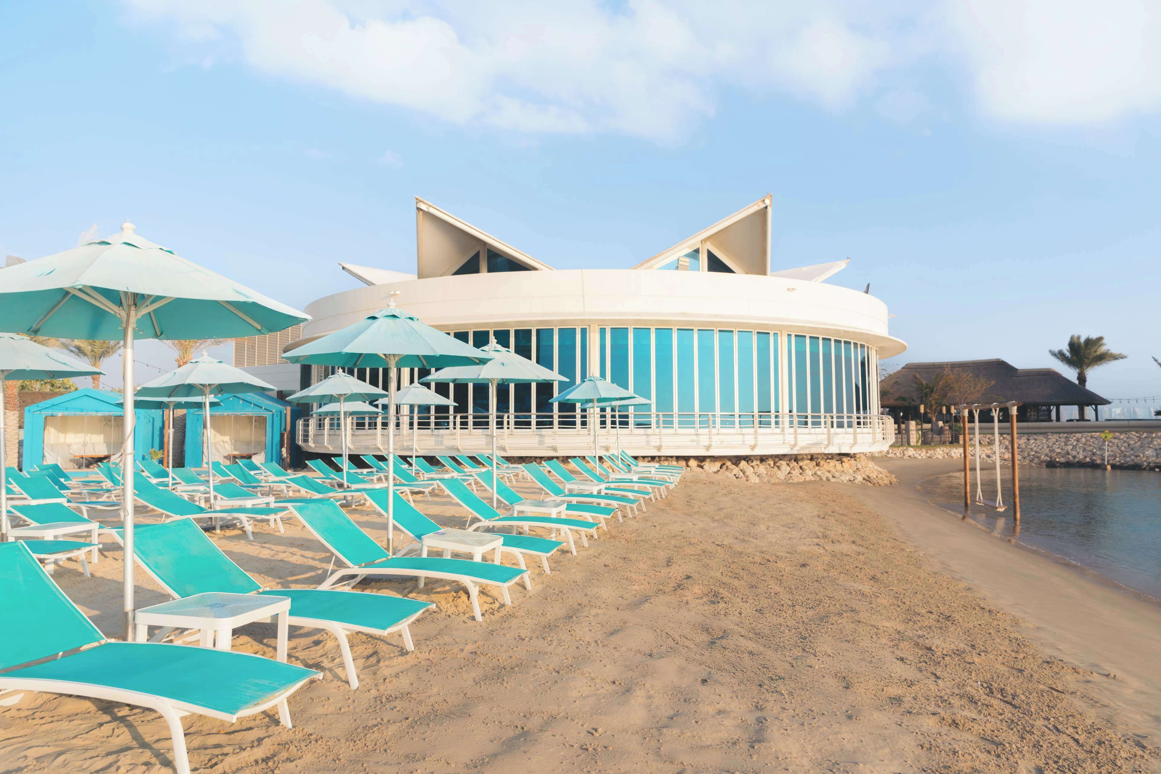 A beachside hotel with a circular building, rows of blue lounge chairs, and umbrellas.