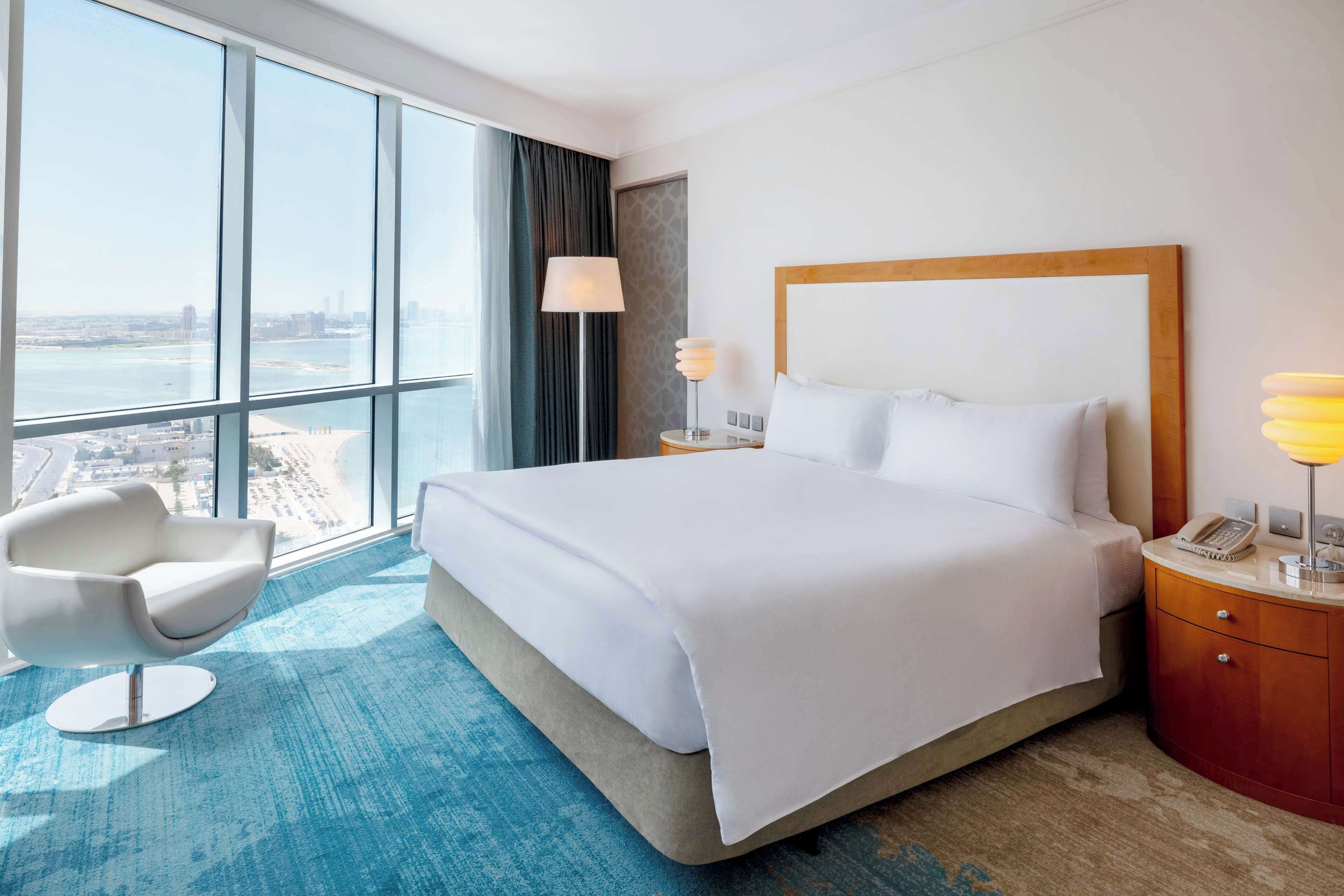 A hotel room with a large bed, a modern chair, a nightstand with a lamp, and a view of the city through large windows.