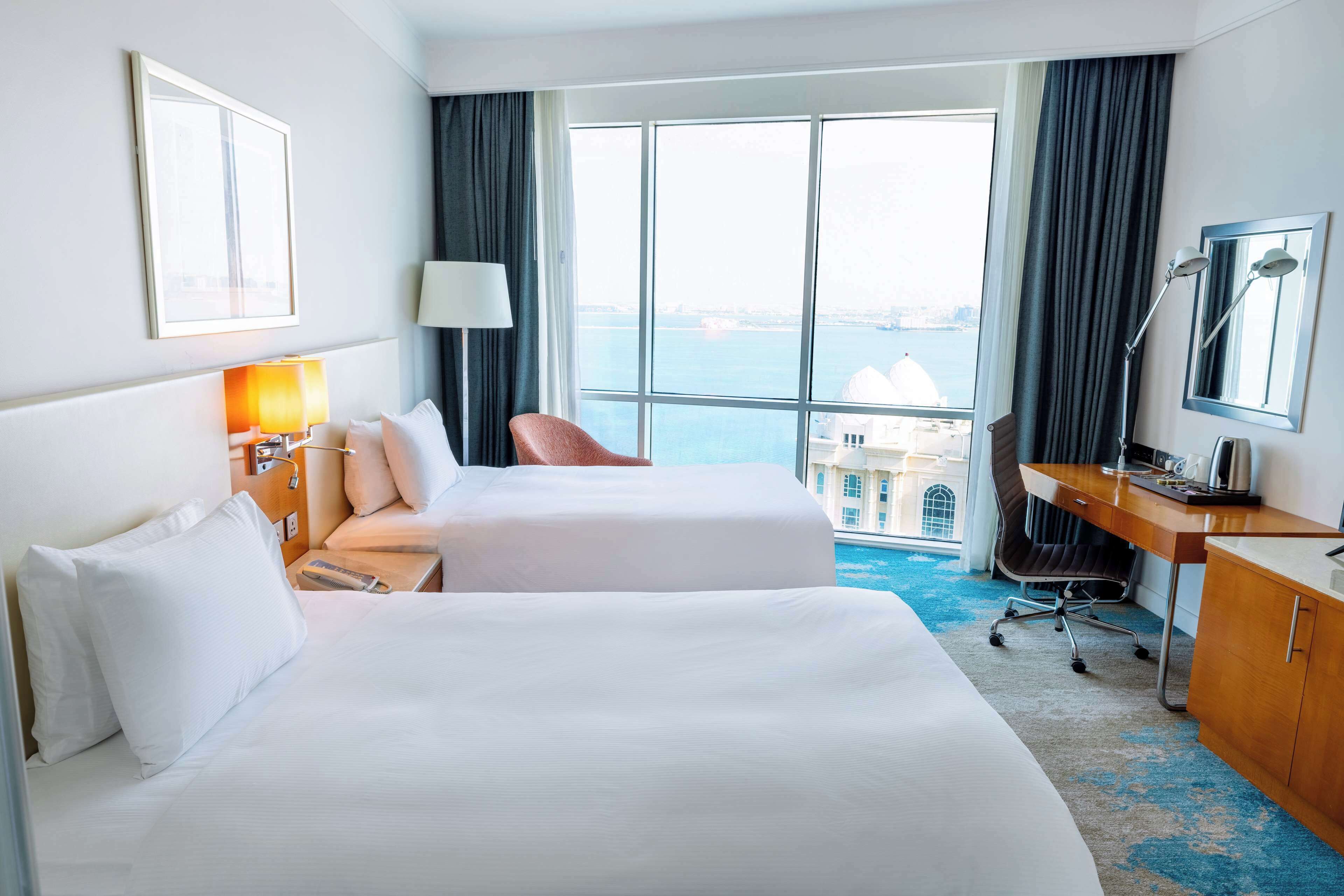 A hotel room with two beds, a desk, a chair, a lamp, and a view of the sea.