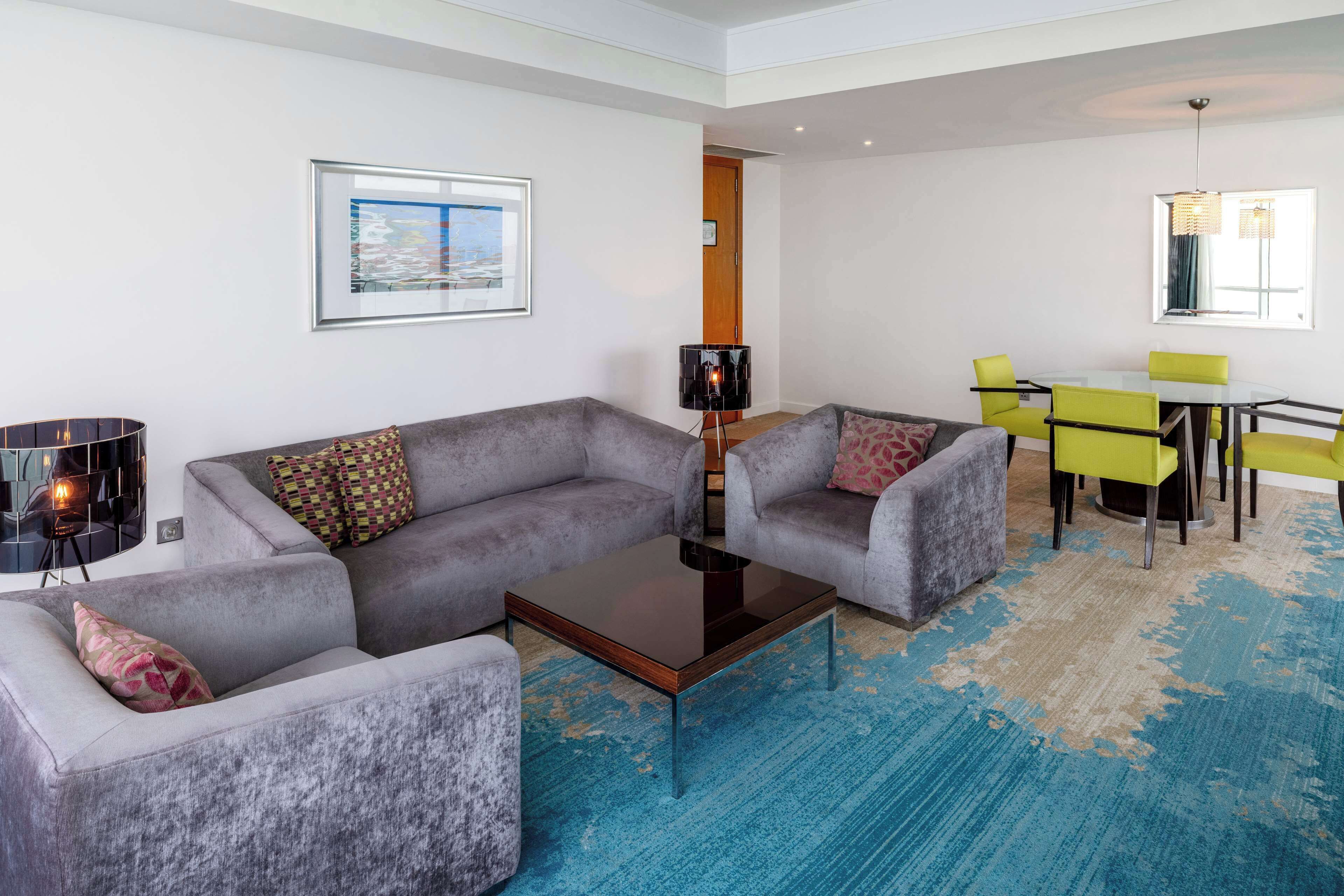 A modern living room with grey sofas, a glass coffee table, and a dining area with yellow chairs.