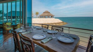 Un comedor con una mesa preparada para cuatro, con vista a una playa y una estructura de techo de paja.