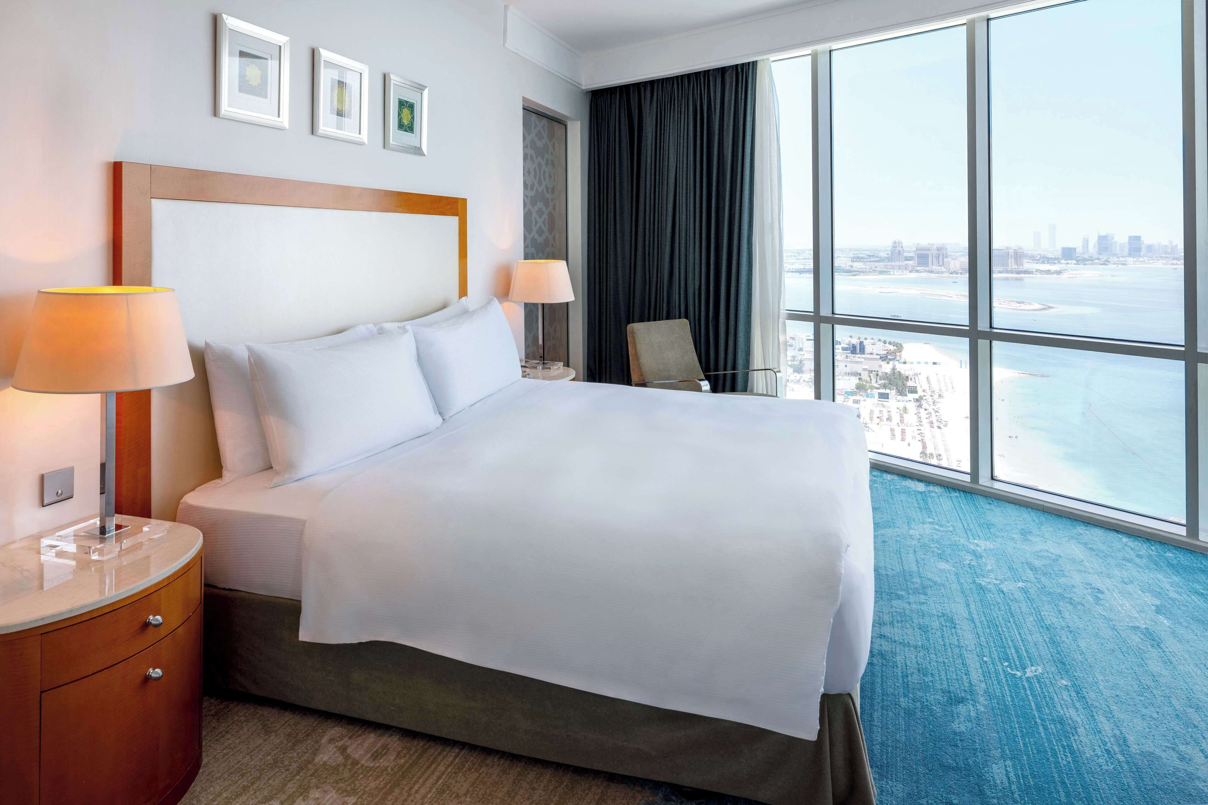 A hotel room with a large bed, two bedside lamps, a nightstand, a chair, and a view of the city and water through large windows.