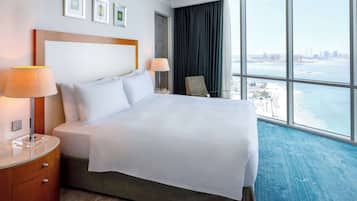 A hotel room with a large bed, two bedside lamps, a nightstand, a chair, and a view of the city and water through large windows.